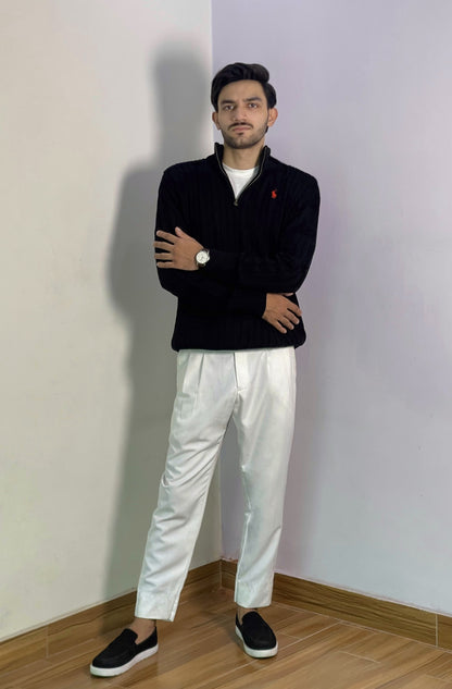 Man wearing a black sweater, white pants, and black shoes standing against a plain wall.