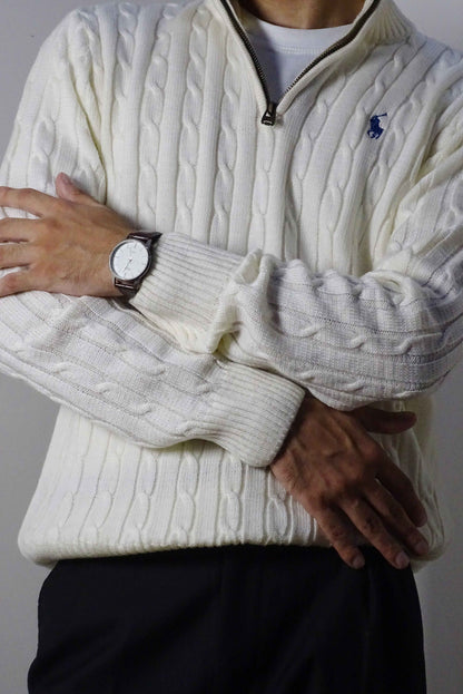Person wearing a white cable knit sweater with a brand logo, arms crossed.