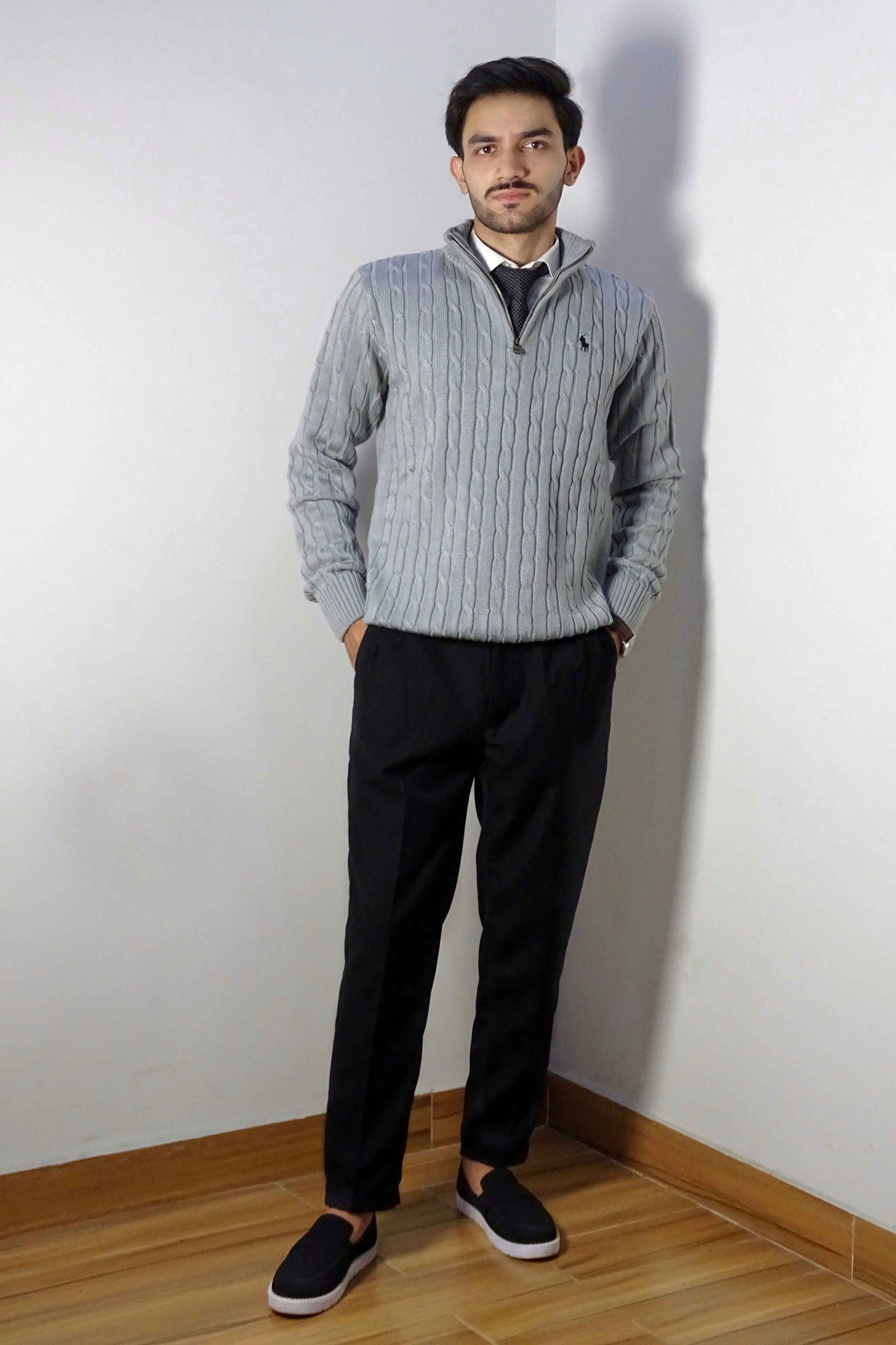 Man wearing a gray sweater and black pants standing against a white wall.