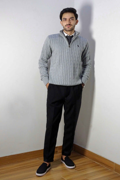 Man wearing a gray sweater and black pants standing against a white wall.
