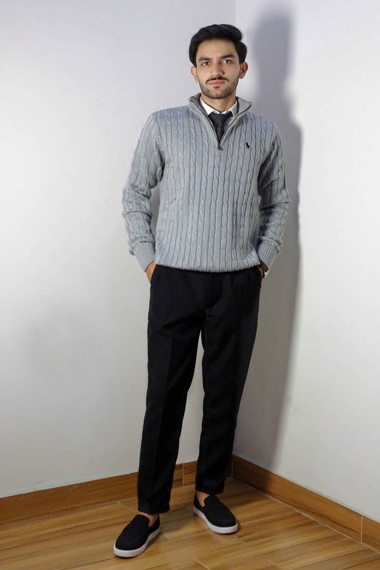 Man wearing a gray sweater and black pants standing against a white wall.