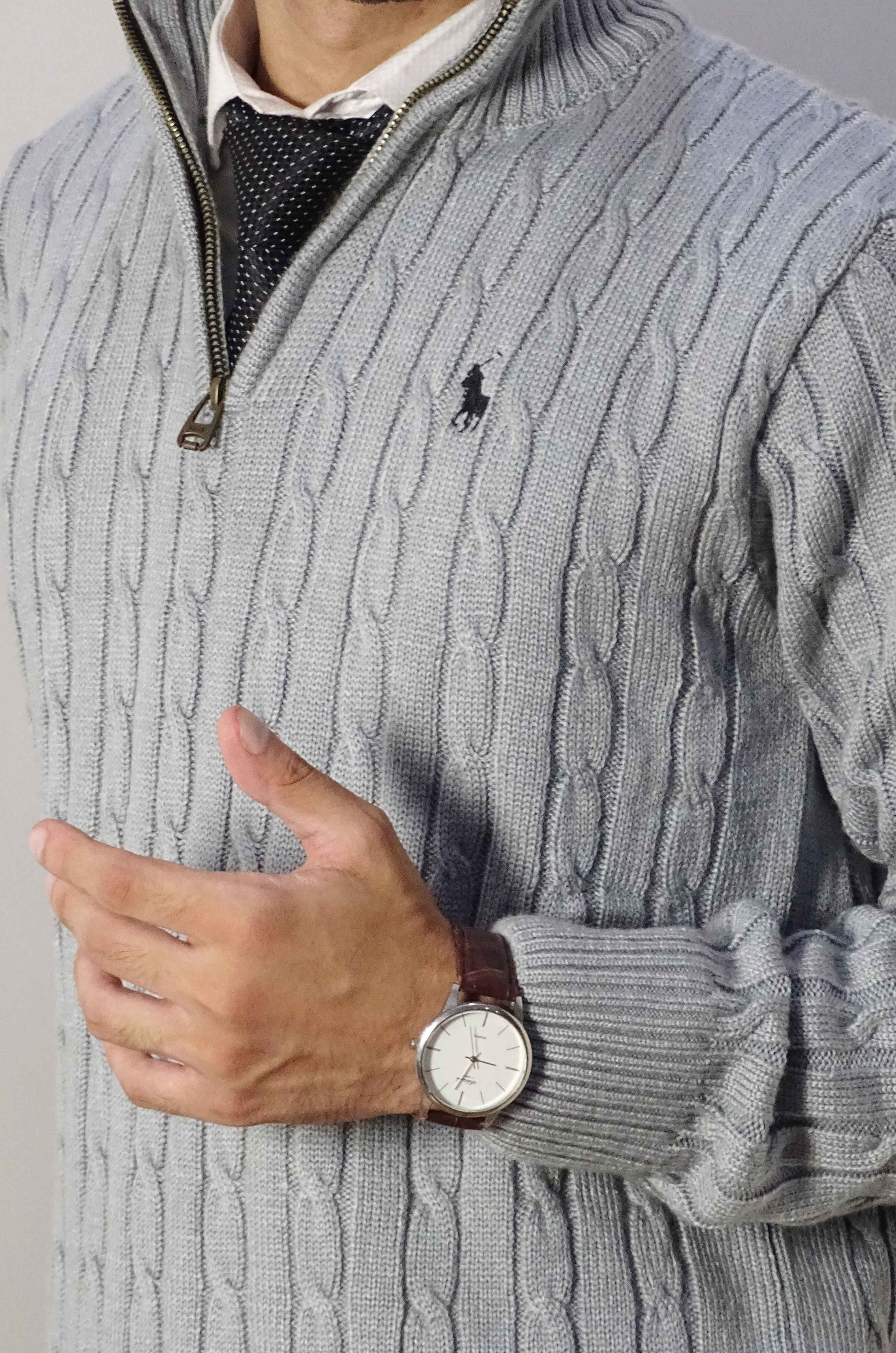 Person wearing a gray cable knit sweater with a brand logo, holding a wristwatch.