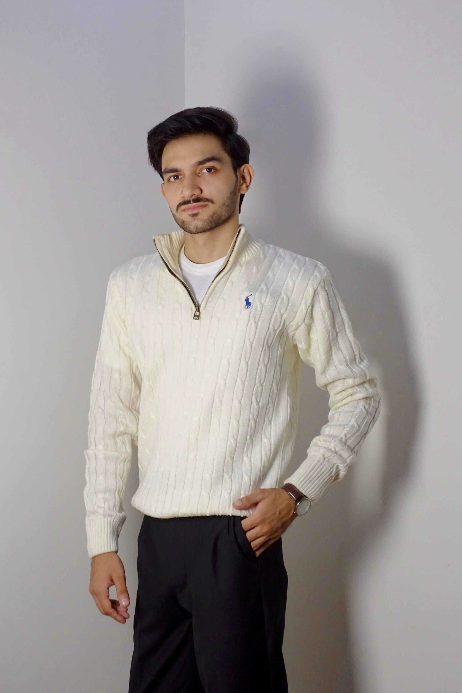 Man wearing a cream-colored cable knit sweater with a brand logo, standing against a plain background.