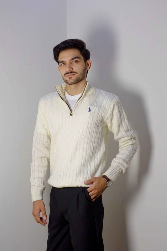 Man wearing a cream-colored cable knit sweater with a brand logo, standing against a plain background.