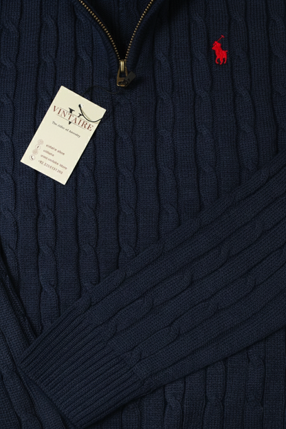Navy cable knit sweater with a red logo tag on a dark background