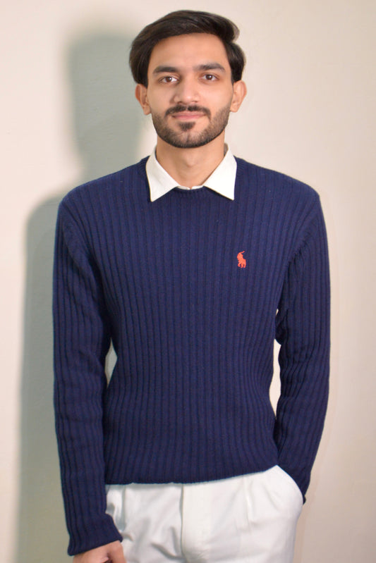Man wearing a navy blue sweater with a red logo over a white shirt, standing against a plain background.