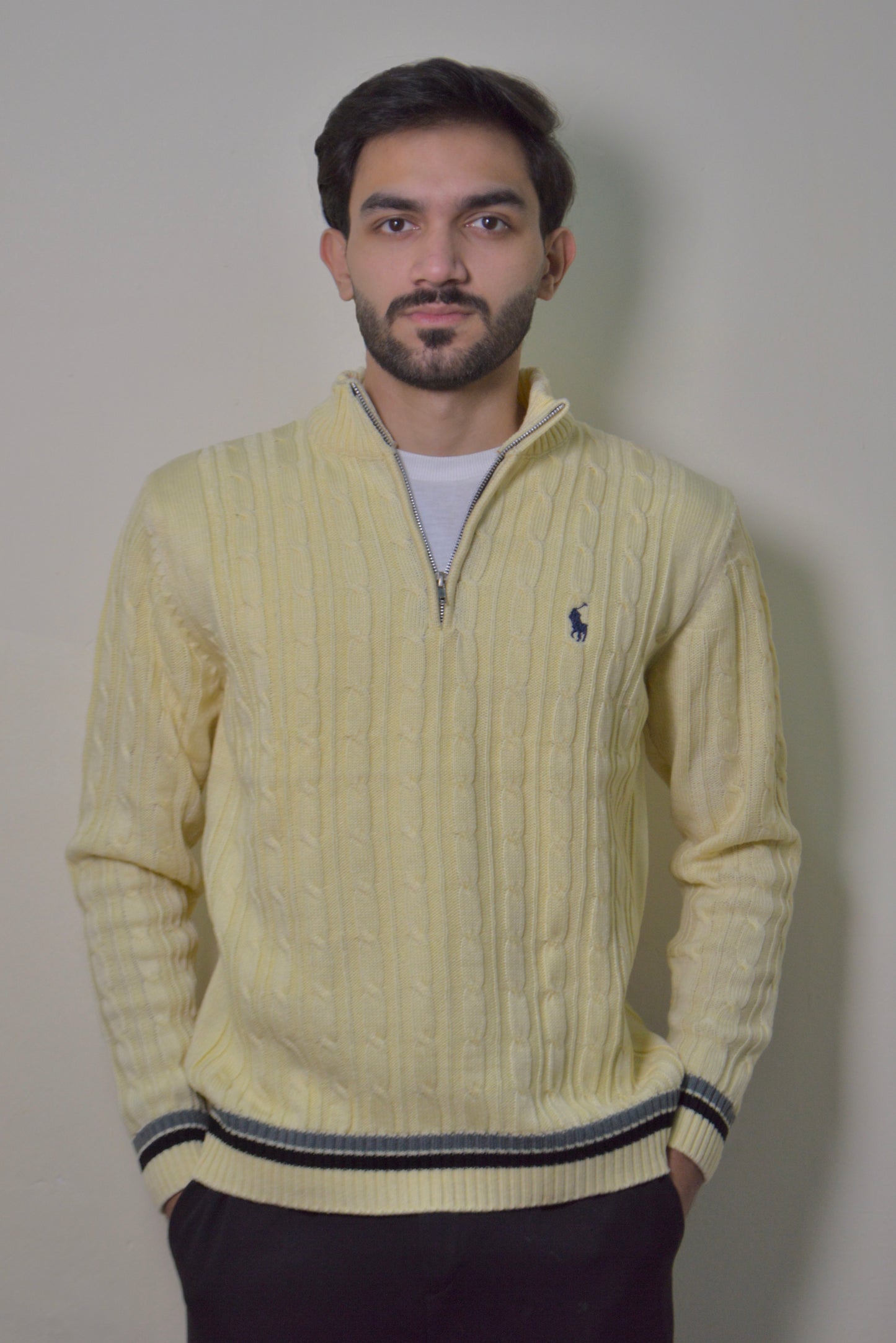 Man wearing a light yellow cable knit sweater with a brand logo against a plain background