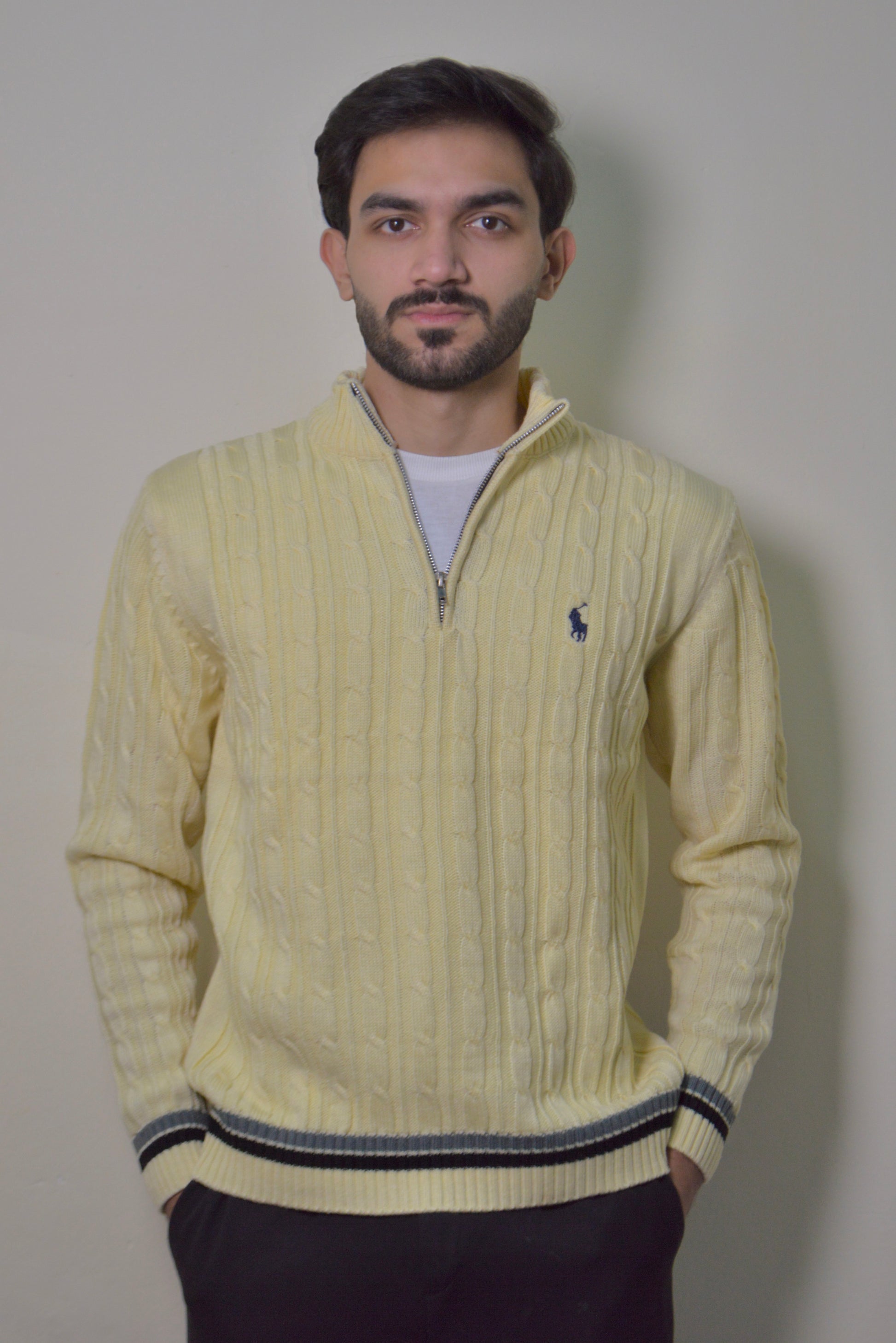 Man wearing a light yellow cable knit sweater with a brand logo against a plain background