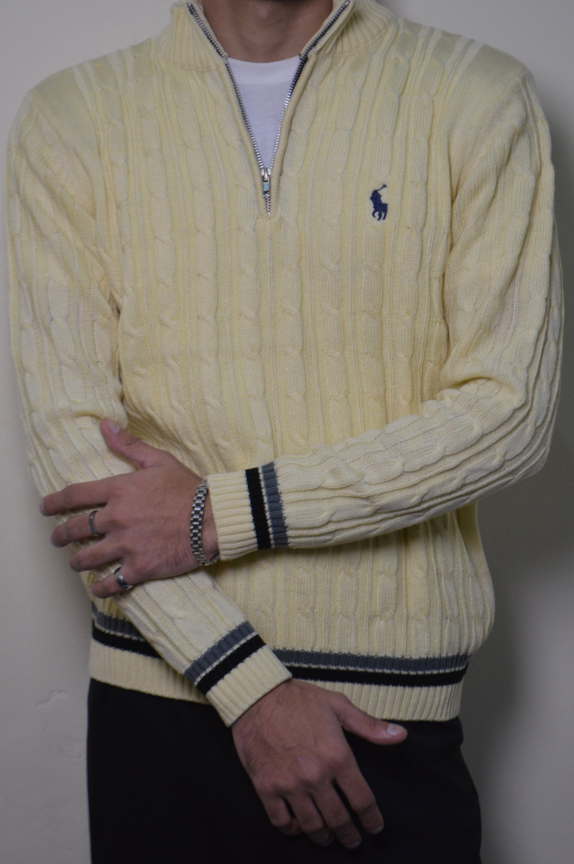 Person wearing a cream-colored cable knit sweater with a brand logo on a plain background