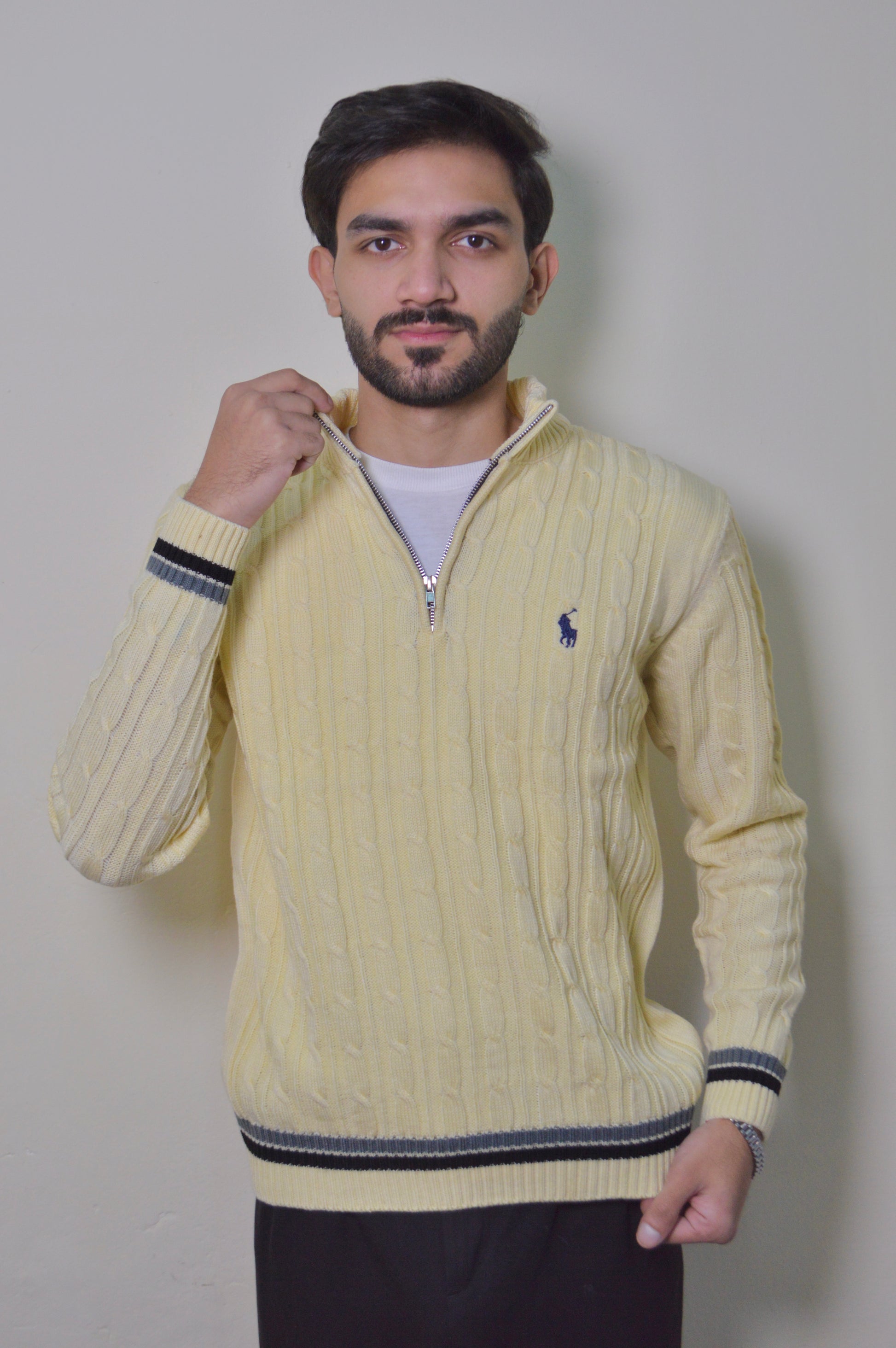 Man wearing a yellow cable knit sweater with a small logo on a plain background